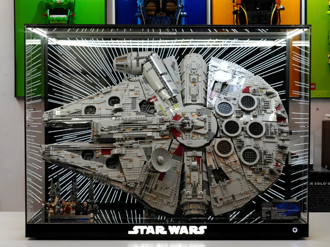 Large LEGO Millennium Falcon model in a glass display case with a starburst background and a row of minifigures at the bottom left; STAR WARS branding at the base.