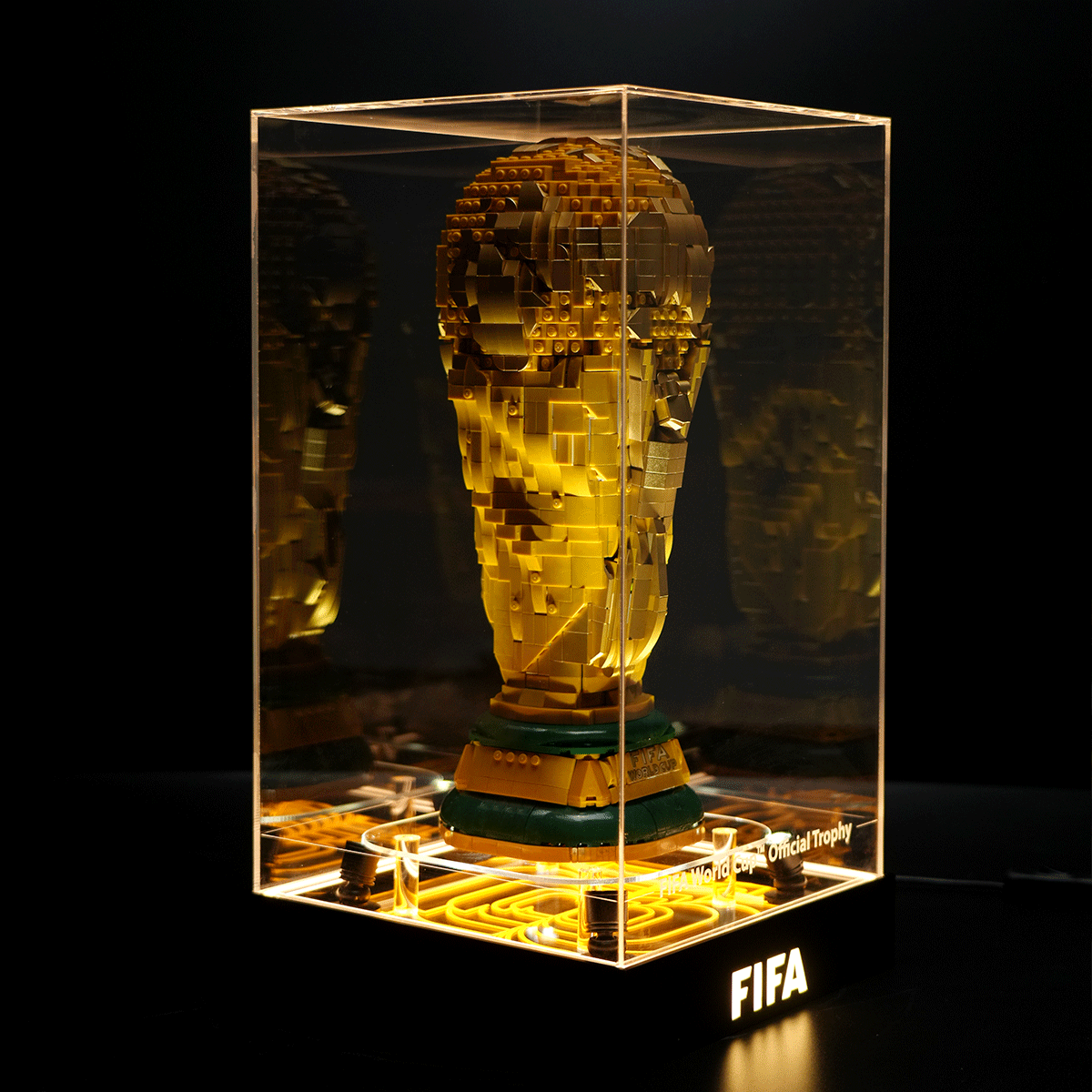 Gold LEGO sculpture of the FIFA World Cup Trophy inside a glass display case, illuminated from underneath against a dark background, with 