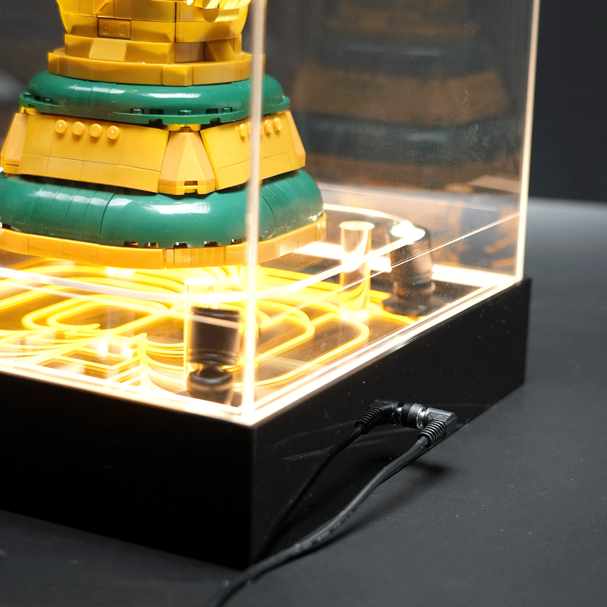 Colorful tiered sculpture with yellow and green rings inside a glass display case, lit by glowing yellow cables at the base.