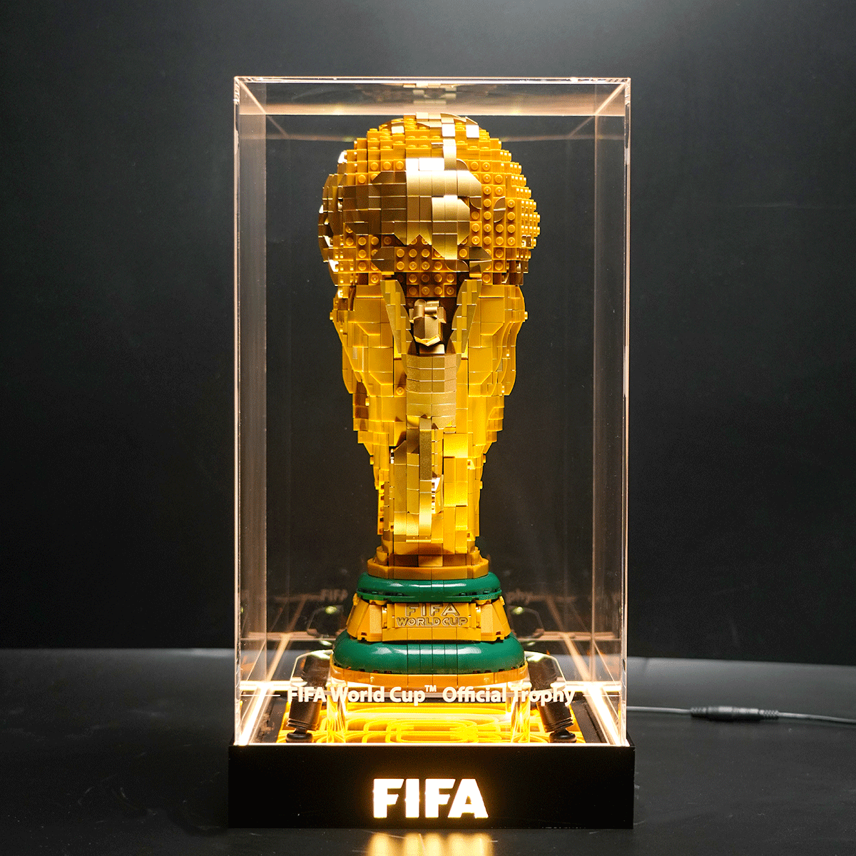 Lego sculpture of the FIFA World Cup trophy inside a clear display case, lit from below, on a dark background.