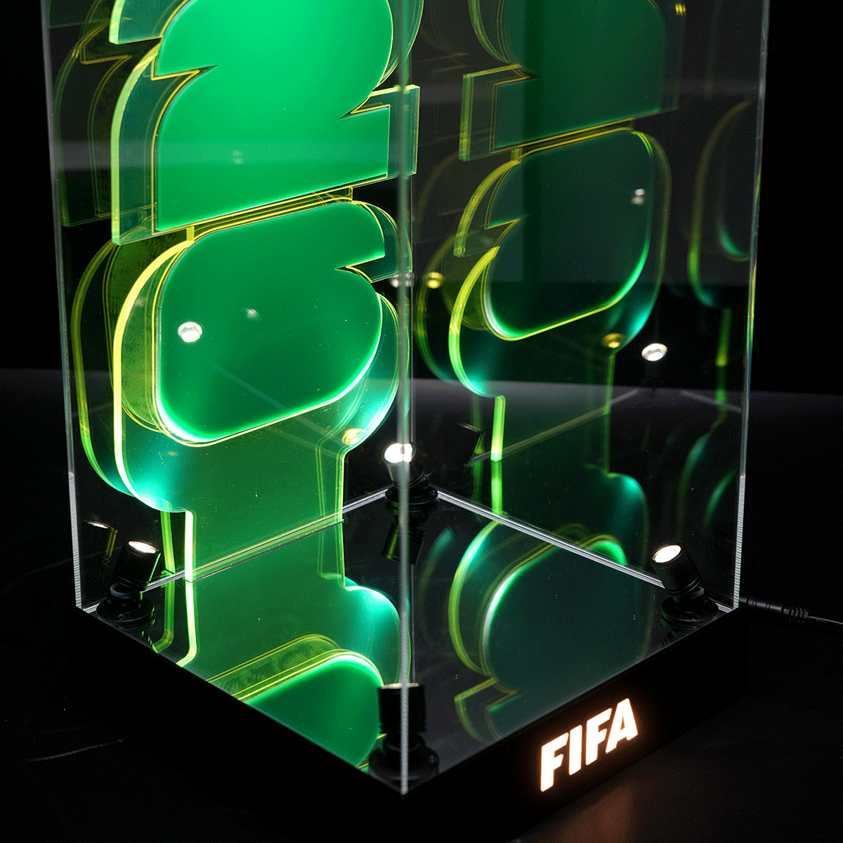 Clear acrylic display cube with oversized green FIFA letters and neon lighting, sitting on a black base with a FIFA logo illuminated at the bottom.