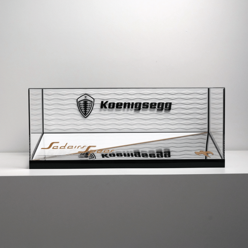 Glass display case with black frame on a white shelf showing a Koenigsegg logo and brand name on the back panel.