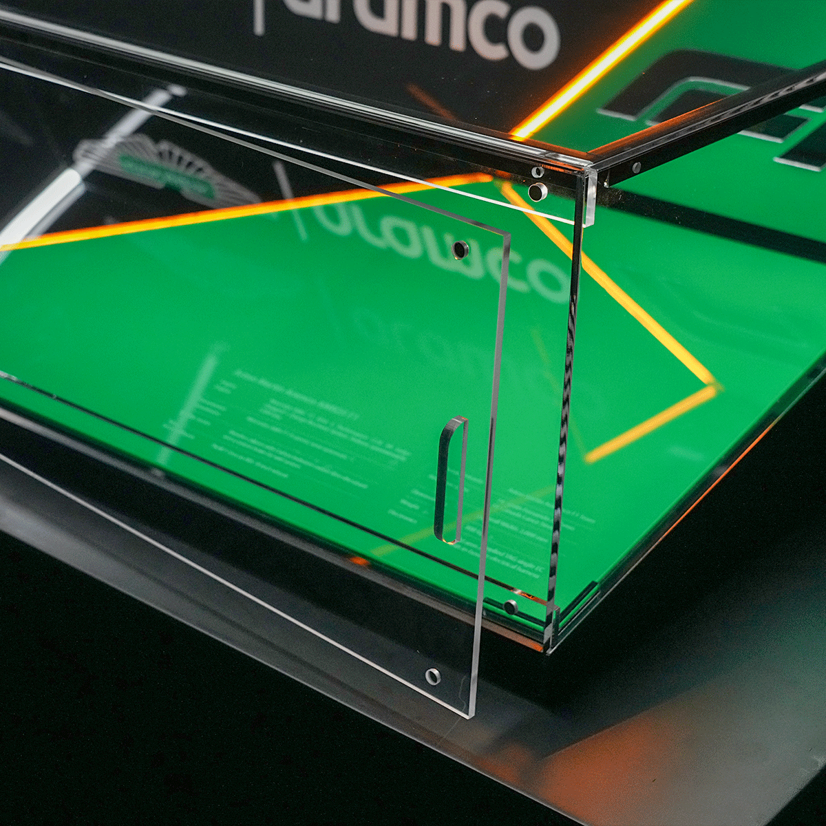 Close-up of a clear glass panel with a slim rectangular cutout, mounted on a metal frame over a bright green display surface with orange neon lines reflecting off the glass.