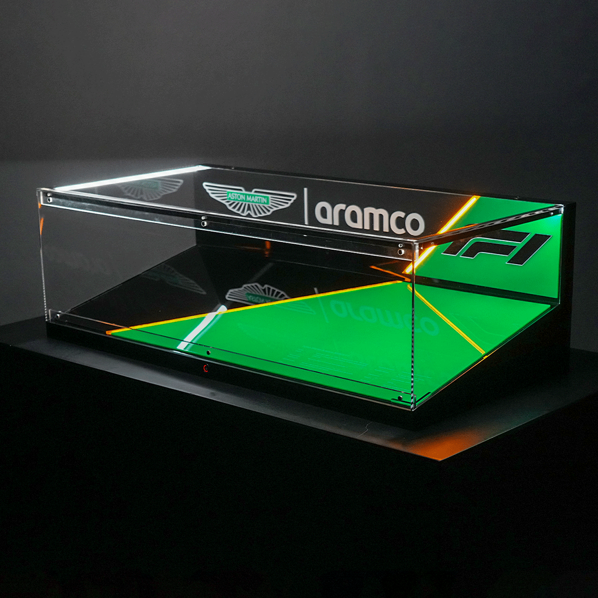 Clear acrylic display case with the Aston Martin Aramco logo and green lighting on a black pedestal.