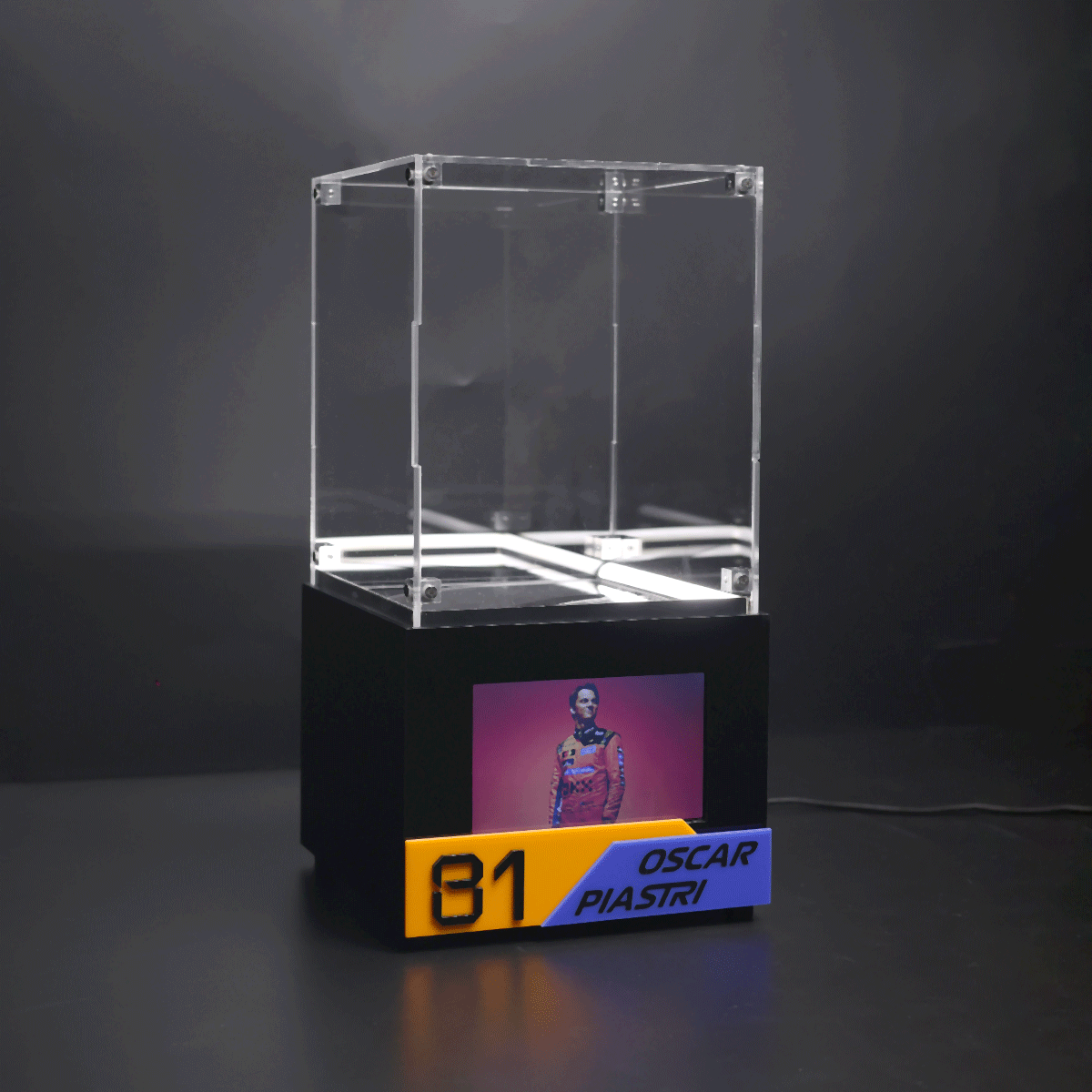 Transparent acrylic display case atop a black pedestal, featuring a poster of a driver and a yellow-blue plate reading '31 Oscar Piastri'.