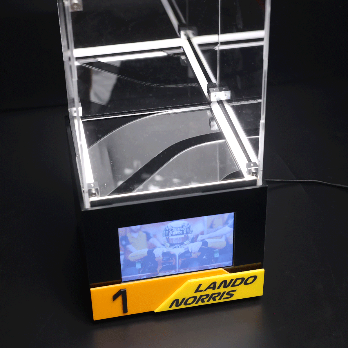 Clear acrylic display case with LED lighting on a black base, showing a small screen and a yellow name plate reading 'LANDO NORRIS' and '1'
