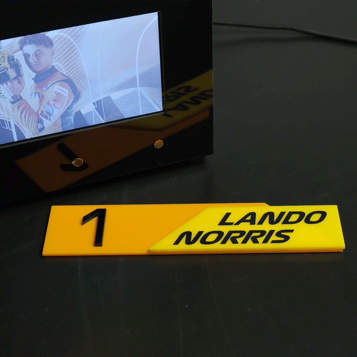 Yellow first-place plaque with the name LANDO NORRIS and the number 1 on a dark table near a monitor displaying a racing driver.