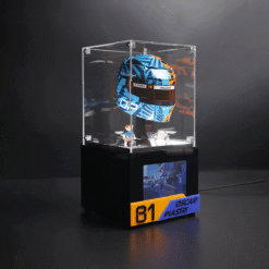 Colorful racing helmet inside a clear acrylic display case on a black pedestal, with a yellow/blue 'Oscar Piastri 81' plaque.