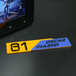 Racing nameplate showing number 81 and the name Oscar Piastri in blue and yellow on a dark desk near a monitor.