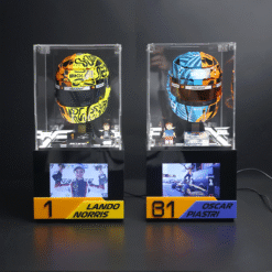Two clear display cases show colorful racing helmets (yellow/black and blue/orange) with mini figurines and podium plaques reading 'Lando Norris' and 'Oscar Piastri' respectively, on a black base with screens beneath.