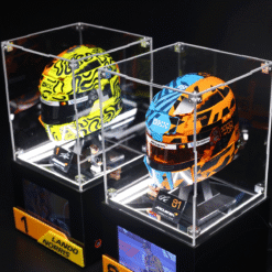 Two colorful racing helmets displayed in clear glass cases on a black pedestal, McLaren-branded and part of a showroom/museum exhibit.