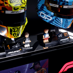 Two LEGO minifigures stand on a display podium beside large colorful McLaren Formula 1 helmets and sponsorship signage.