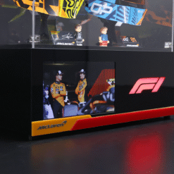 McLaren F1 display: toy driver figures and model cars on a glass shelf above a screen showing two drivers in orange racing suits.