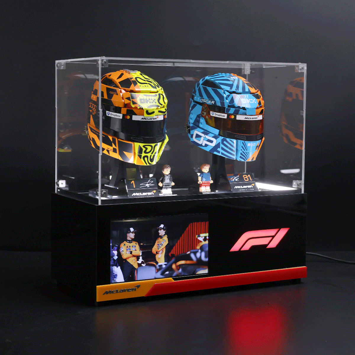 Two colorful Formula 1 helmets in a clear glass display case on a black pedestal,McLaren branding visible below.