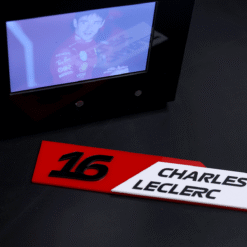 Close-up of a red number plate '16' beside a white nameplate reading 'CHARLES LECLERC' on a dark surface, with a small screen showing a blue-tinted image of a race driver in a red suit above them.