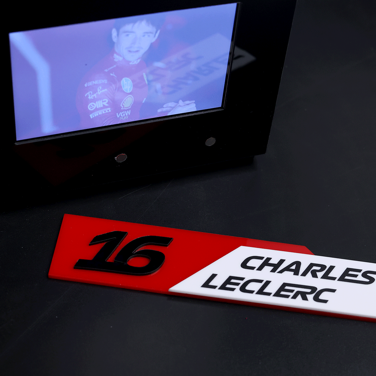 Close-up of a red number plate '16' beside a white nameplate reading 'CHARLES LECLERC' on a dark surface, with a small screen showing a blue-tinted image of a race driver in a red suit above them.