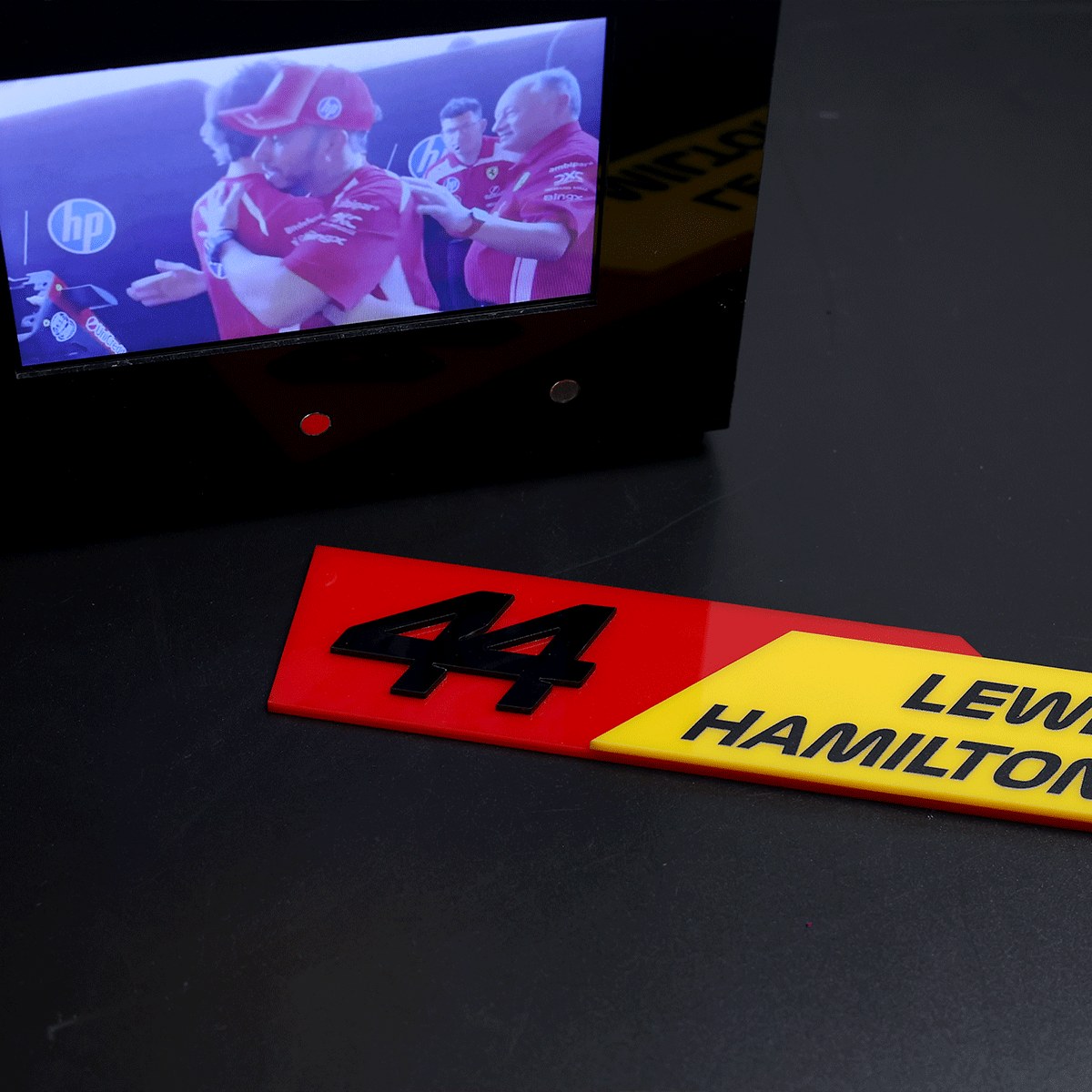 Close-up of a red race plate with the number 44 and a yellow Lewis Hamilton plate beside a monitor showing a team in red uniforms.