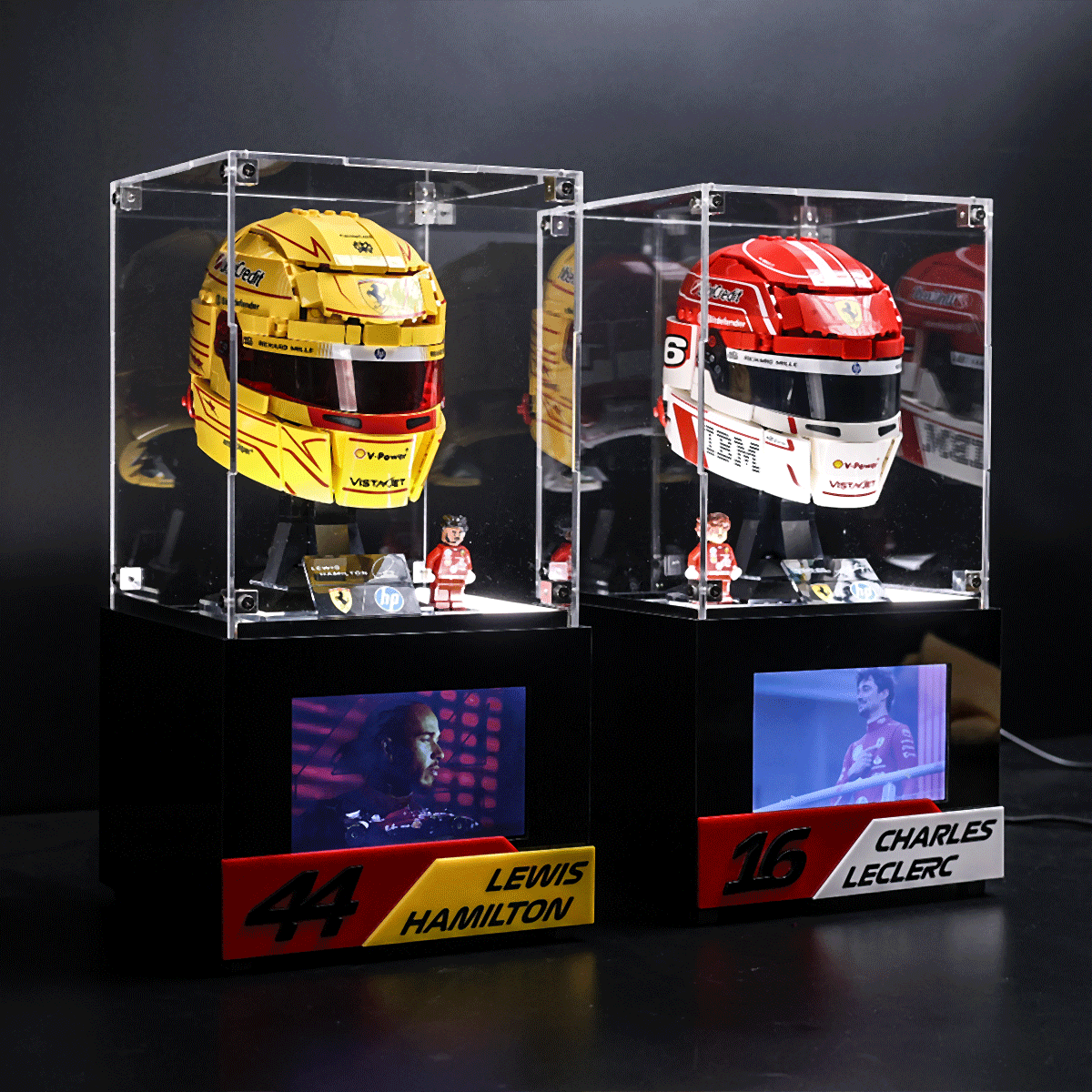 Two race helmet collectibles in glass cases on black stands, labeled Lewis Hamilton (left) and Charles Leclerc (right).