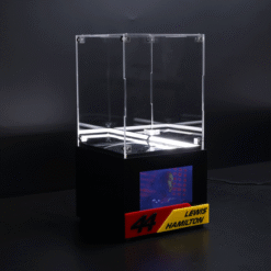 Clear acrylic display case atop a black pedestal, featuring a small screen showing Lewis Hamilton and a red‑yellow name plaque reading '44 Lewis Hamilton'