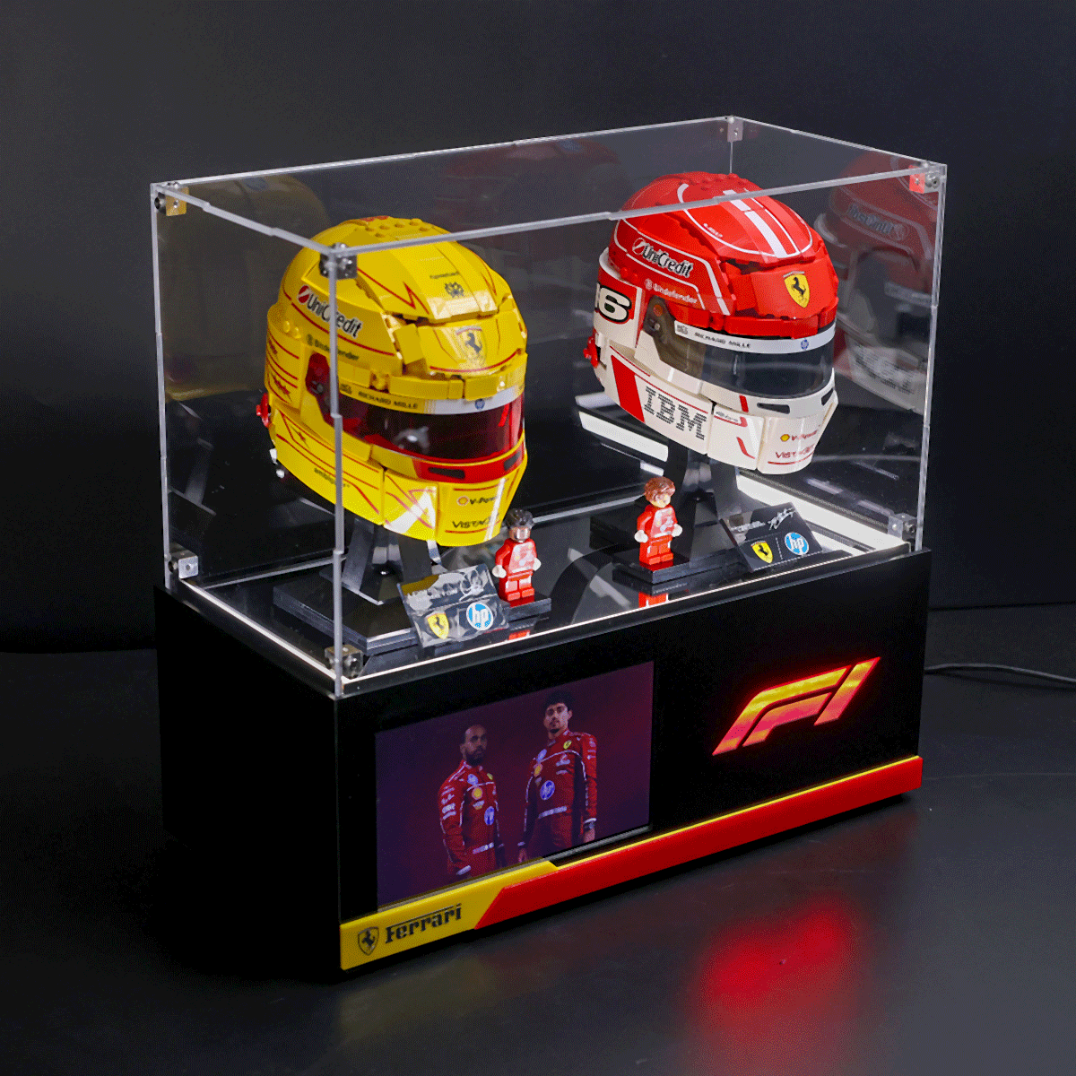 Glass display case featuring yellow and red Ferrari Formula 1 helmets with two small figurines, on a black pedestal with Ferrari branding.