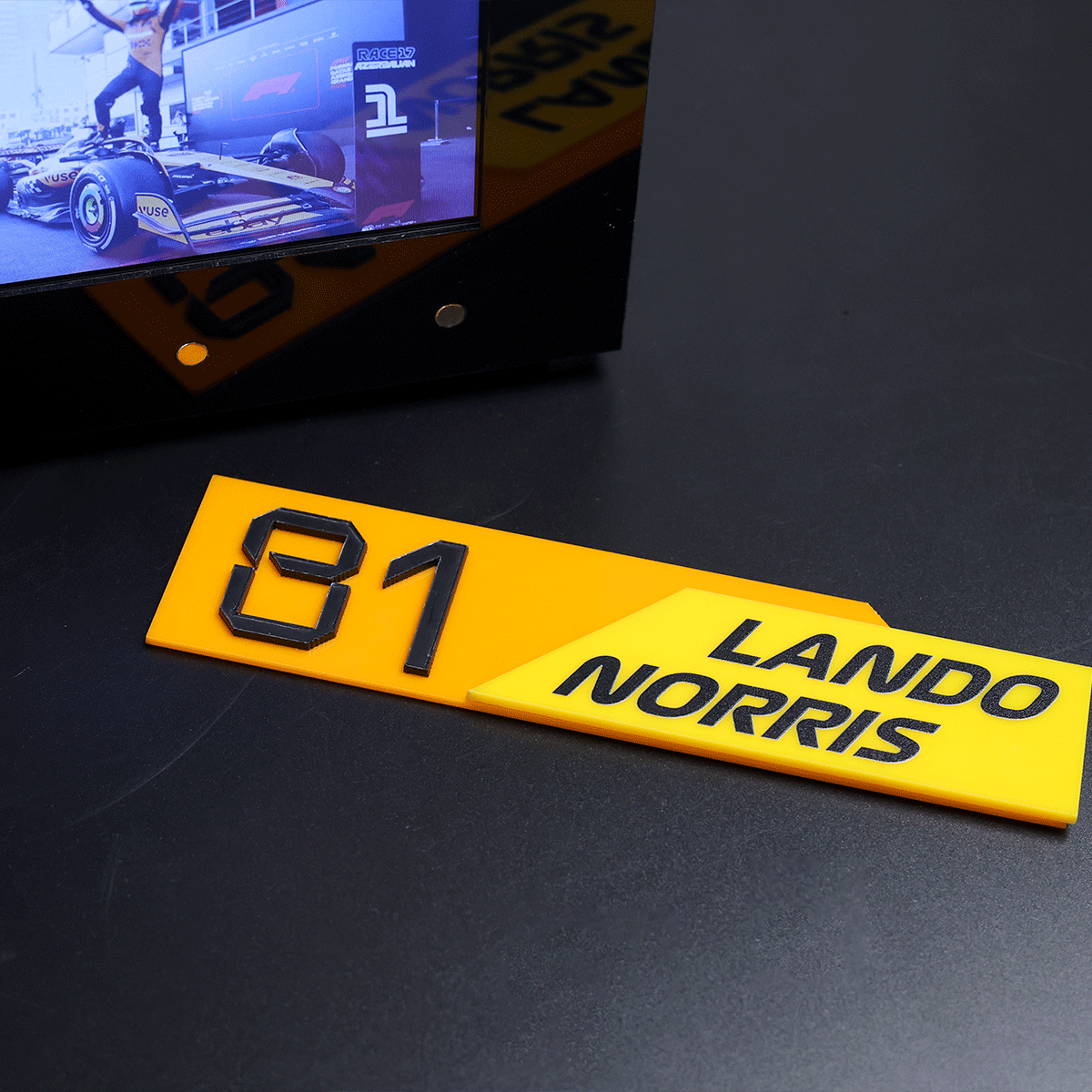 Yellow race placards reading '81' and 'Lando Norris' on a dark table surface, with a monitor in the background.