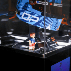 Lego minifigure stands on a display pedestal beside a large blue race car model with sponsor stickers in a glass case.