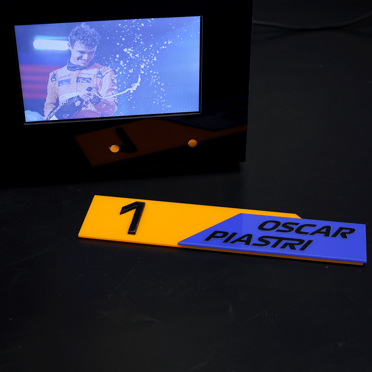 Yellow first-place plaque with a bold 1 sits in front of a blue OSCAR PIASTRI label; a screen shows a driver spraying champagne in celebration.