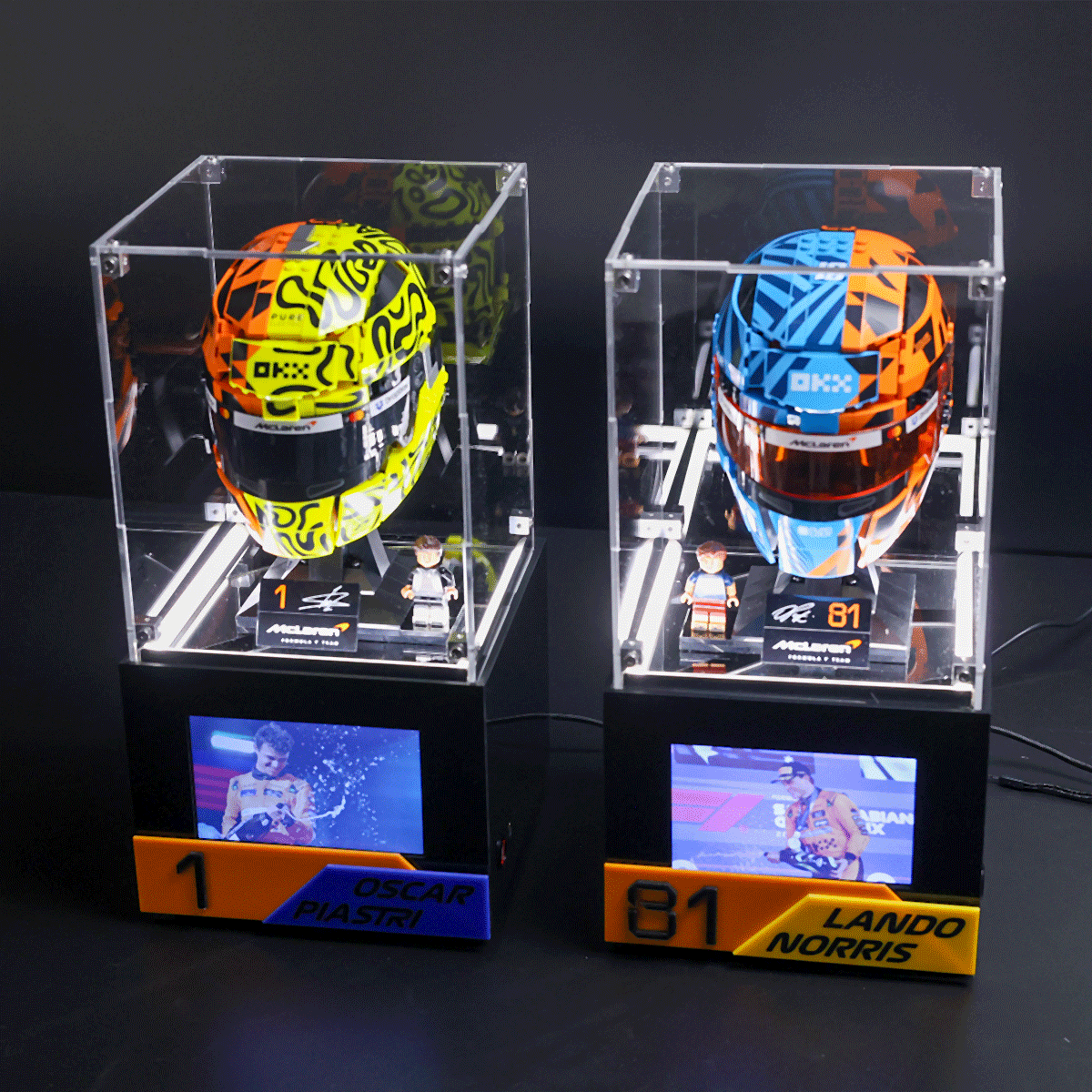 Two brightly colored racing helmets in clear display cases with miniature drivers and nameplates for Oscar Piastri and Lando Norris.