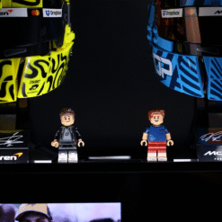 Two LEGO minifigures stand on a black shelf between large yellow and blue racing helmets bearing sponsor logos.