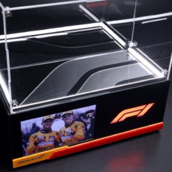 Glass display case with illuminated edges, McLaren logo, and a photo of two Formula 1 drivers in yellow racing suits on a black stand.